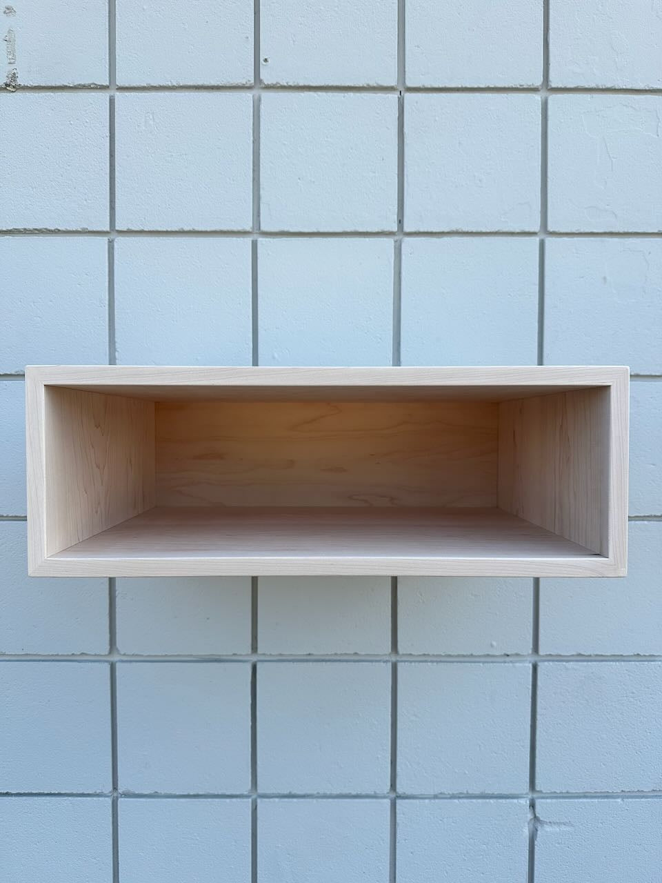 A minimalist, rectangular wooden floating shelf with a light natural finish, mounted on a light blue tiled wall.