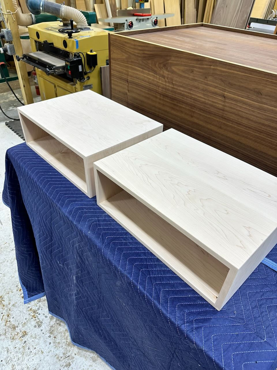 Two unfinished light wood rectangular storage shelves with open cubby sides, placed on a blue quilted moving blanket in a woodworking shop. A yellow woodworking machine and a larger wood panel are visible in the background.