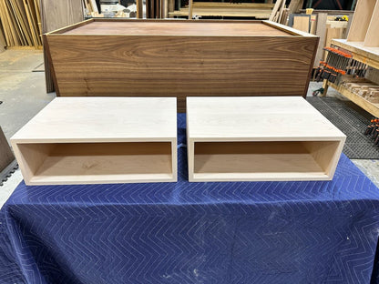 Two unfinished light wood rectangular storage shelves placed side by side on a blue moving blanket, with a larger unfinished wood box behind them in a workshop setting.