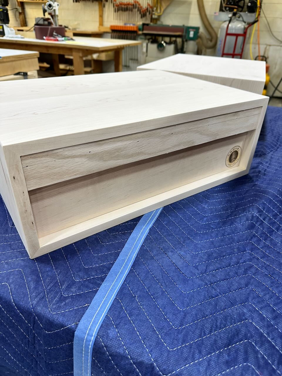 Rear view of wooden nightstand with a recessed panel, resting on a blue quilted moving blanket in a workshop setting.