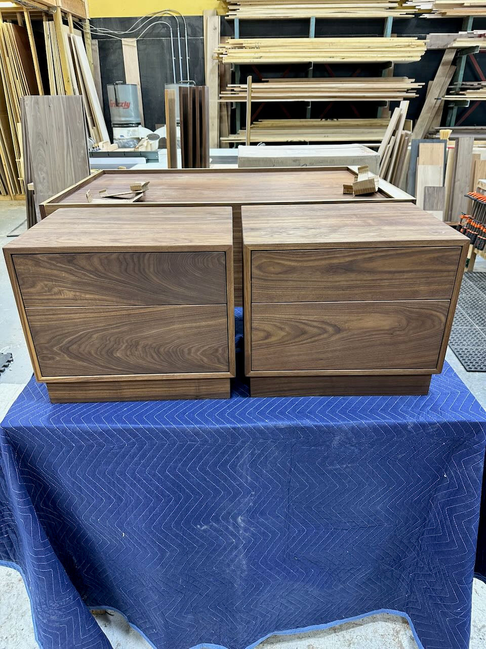 A two-drawer wooden nightstand with a dark walnut finish, resting on a blue moving blanket inside a woodworking shop.