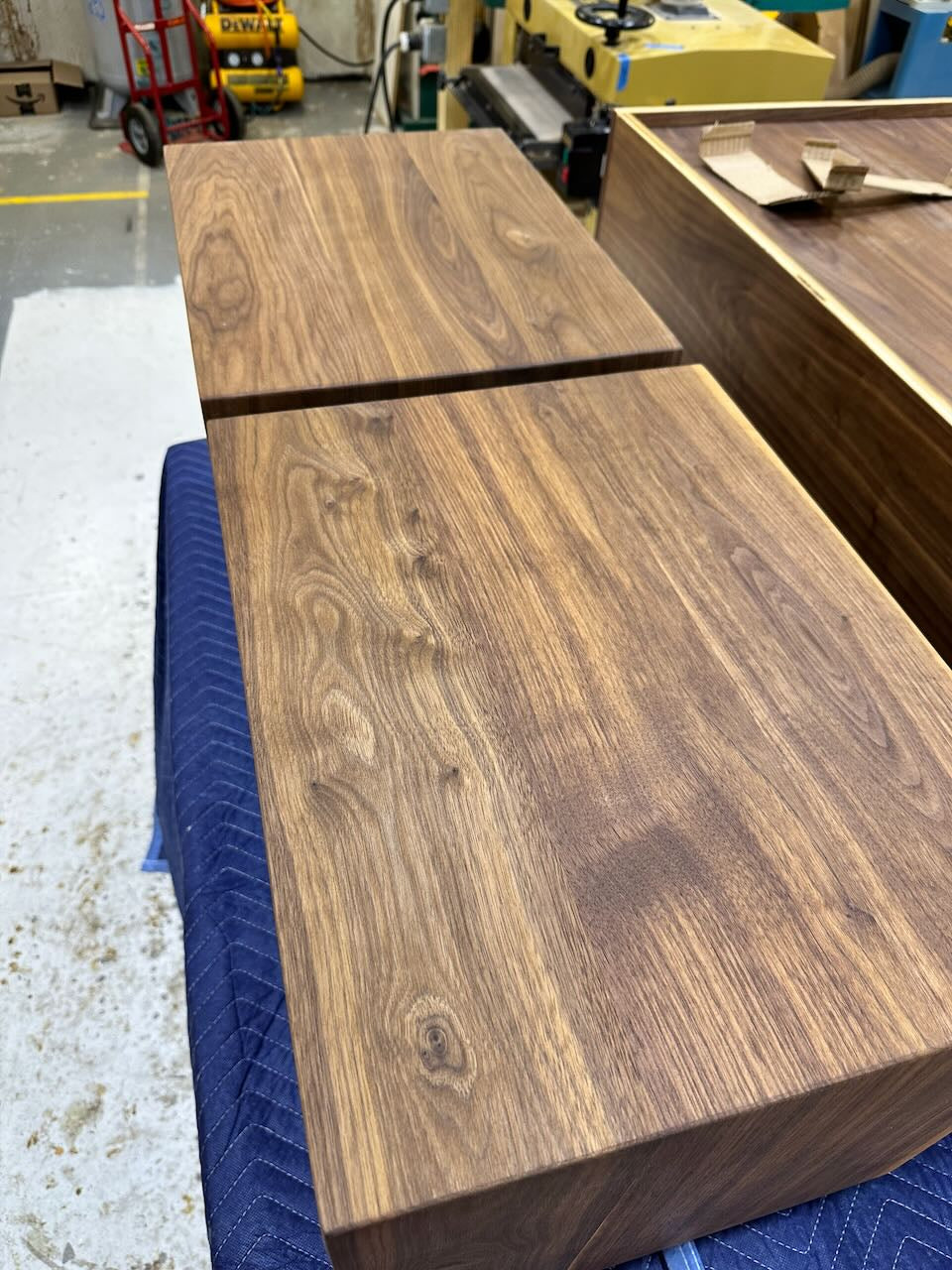 Top views of two wooden nightstands with a rich walnut grain pattern, stacked on a blue quilted surface inside a workshop.