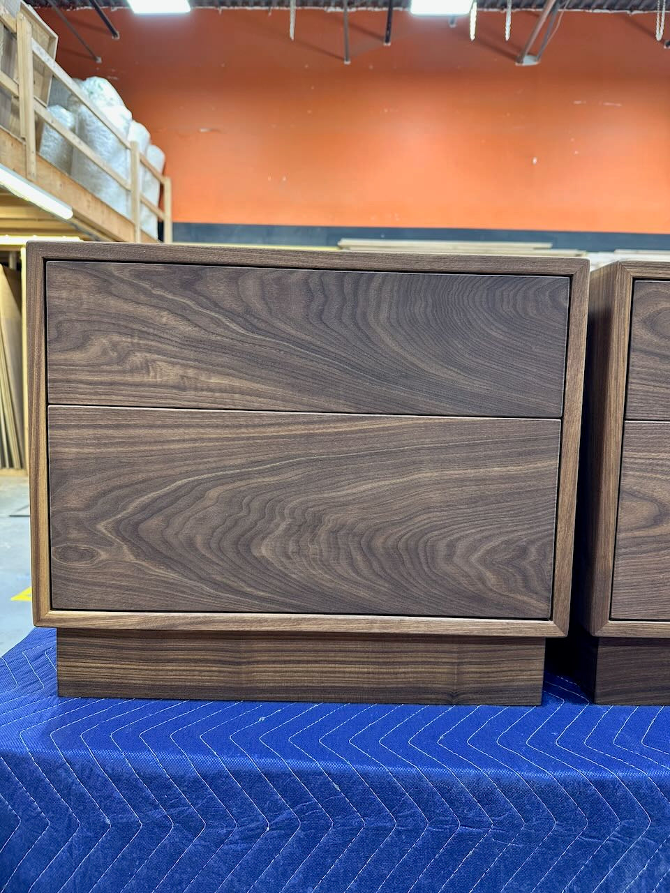 A dark walnut wood two-drawer nightstand with a modern block base, shown alongside another matching nightstand on a blue moving blanket in a warehouse setting.
