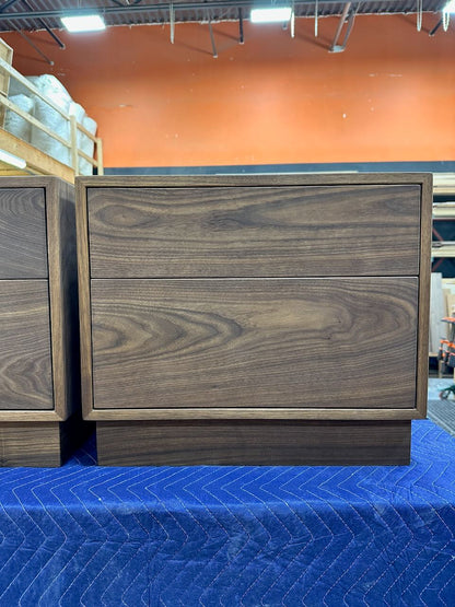 A dark walnut wood two-drawer nightstand with a block base, shown next to another matching nightstand on a blue moving blanket in a warehouse setting.