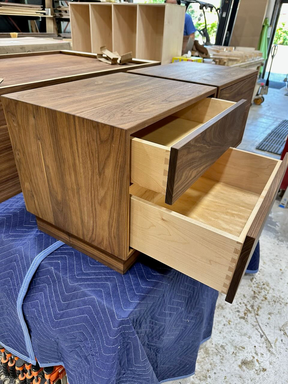 A wooden nightstand with two open drawers, photographed in a workshop setting. The front features a dark walnut wood grain, while the drawer interiors and drawer fronts have a lighter natural wood tone.