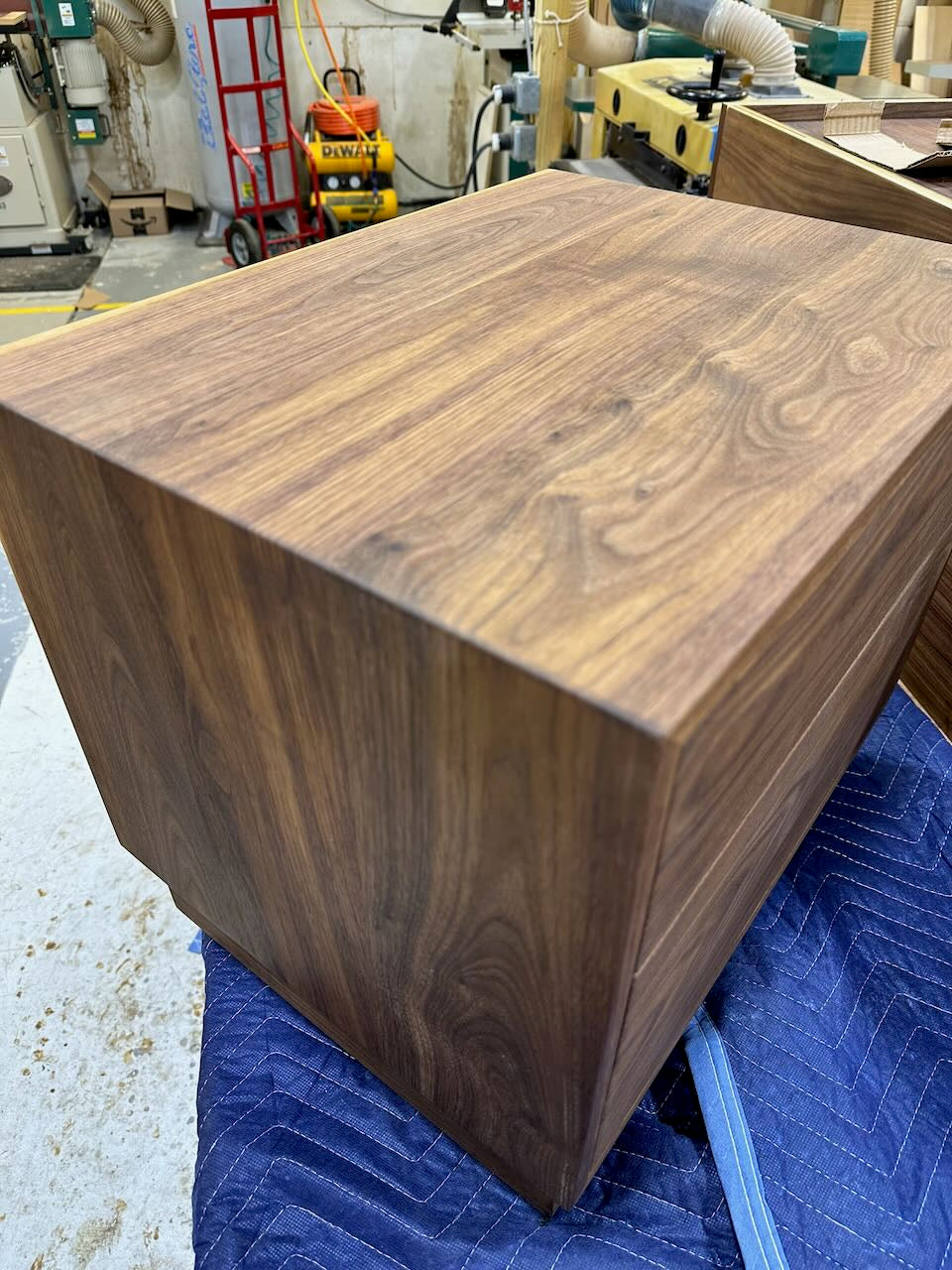 Wooden nightstand with a rich walnut finish, resting on a blue quilted moving blanket inside a workshop environment.