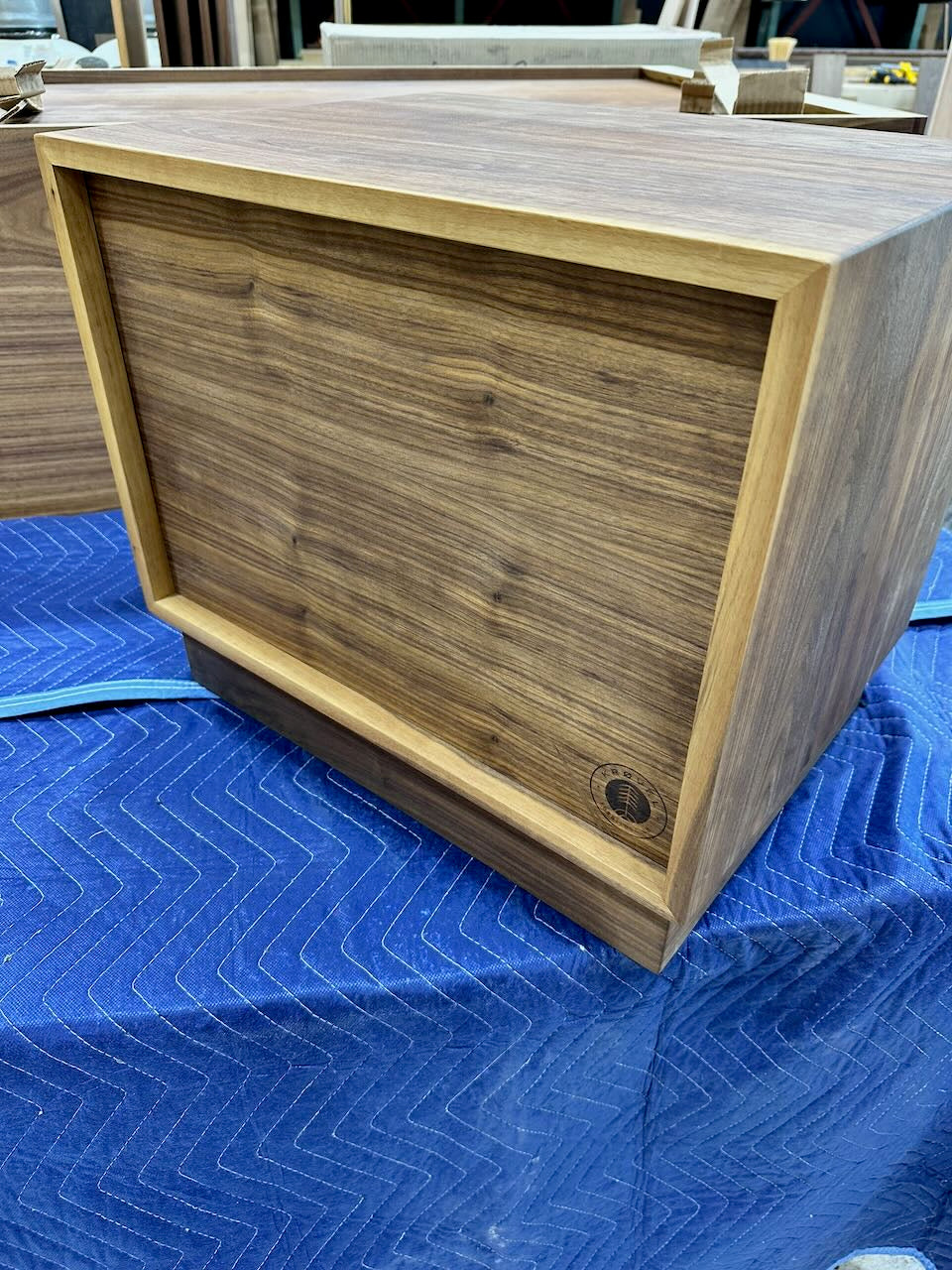 Back of wooden nightstand with a rich walnut-grain finish, resting on a blue moving blanket in what appears to be a workshop or warehouse setting.