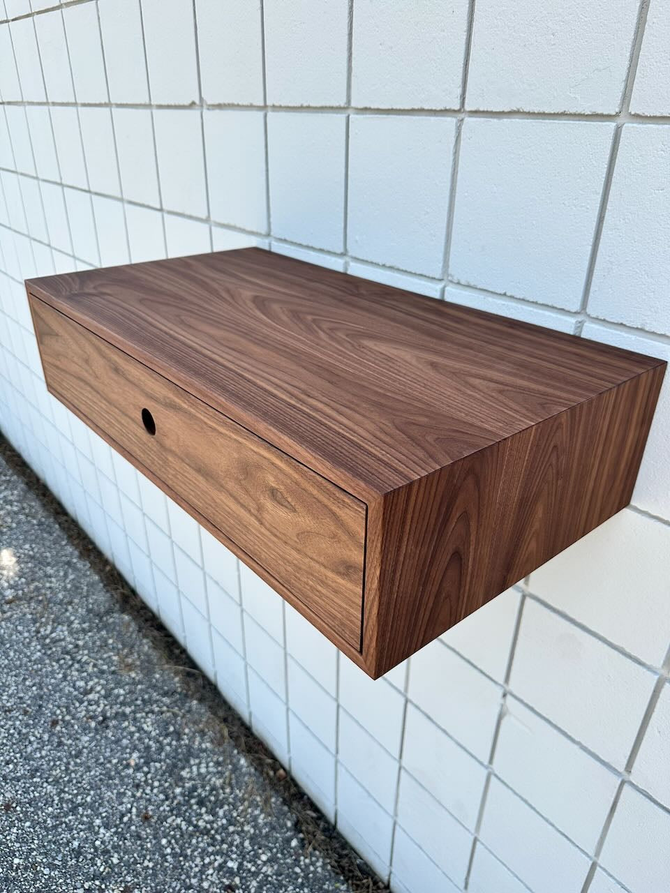 A Krovel Furniture Co. Single Drawer Walnut Desk - SAMPLE, featuring a natural finish and visible wood grain, is mounted on a white tiled wall above a gravel surface. The desk’s single drawer has a round hole handle, evoking the look of solid hardwood.