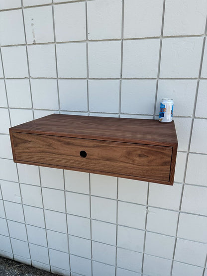 A piece from Krovel Furniture Co., the Single Drawer Walnut Desk - SAMPLE, evokes minimalist elegance with its clean lines and distinctive round hole at the center of the front panel. Mounted on a white tiled wall, this wooden shelf-like desk supports a can of LaCroix sparkling water resting neatly on top.