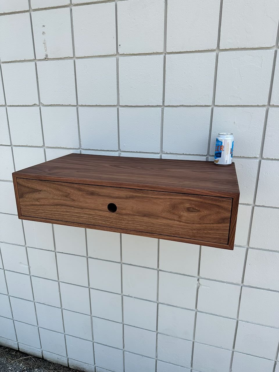 A piece from Krovel Furniture Co., the Single Drawer Walnut Desk - SAMPLE, evokes minimalist elegance with its clean lines and distinctive round hole at the center of the front panel. Mounted on a white tiled wall, this wooden shelf-like desk supports a can of LaCroix sparkling water resting neatly on top.