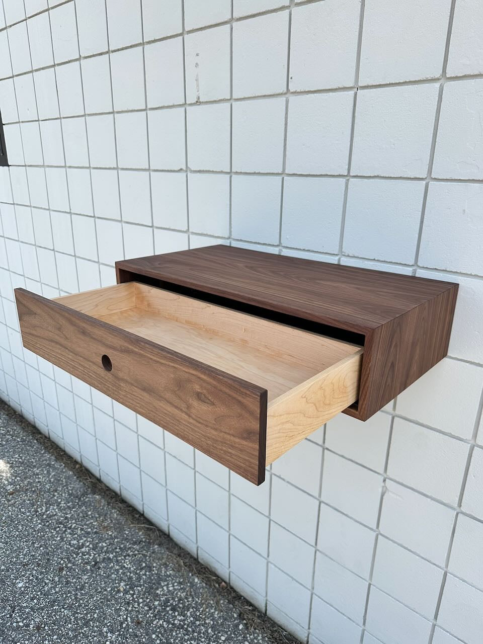 A Krovel Furniture Co. Single Drawer Walnut Desk - SAMPLE with a dark walnut exterior and lighter interior is mounted on a tiled white wall. Its drawer is partially open, showing the empty interior, and gravel ground is visible below.