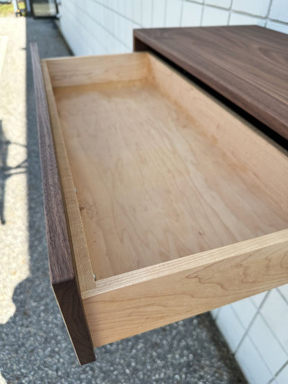 A close-up of the open drawer from the Single Drawer Walnut Desk - SAMPLE by Krovel Furniture Co. reveals its empty light wood interior. This solid hardwood desk, featuring a rich walnut finish, is positioned against a tiled white wall with sunlight streaming in from an outdoor area.