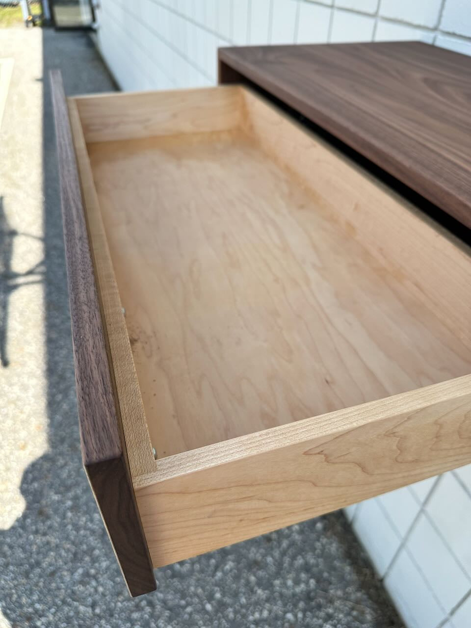 A close-up of the open drawer from the Single Drawer Walnut Desk - SAMPLE by Krovel Furniture Co. reveals its empty light wood interior. This solid hardwood desk, featuring a rich walnut finish, is positioned against a tiled white wall with sunlight streaming in from an outdoor area.