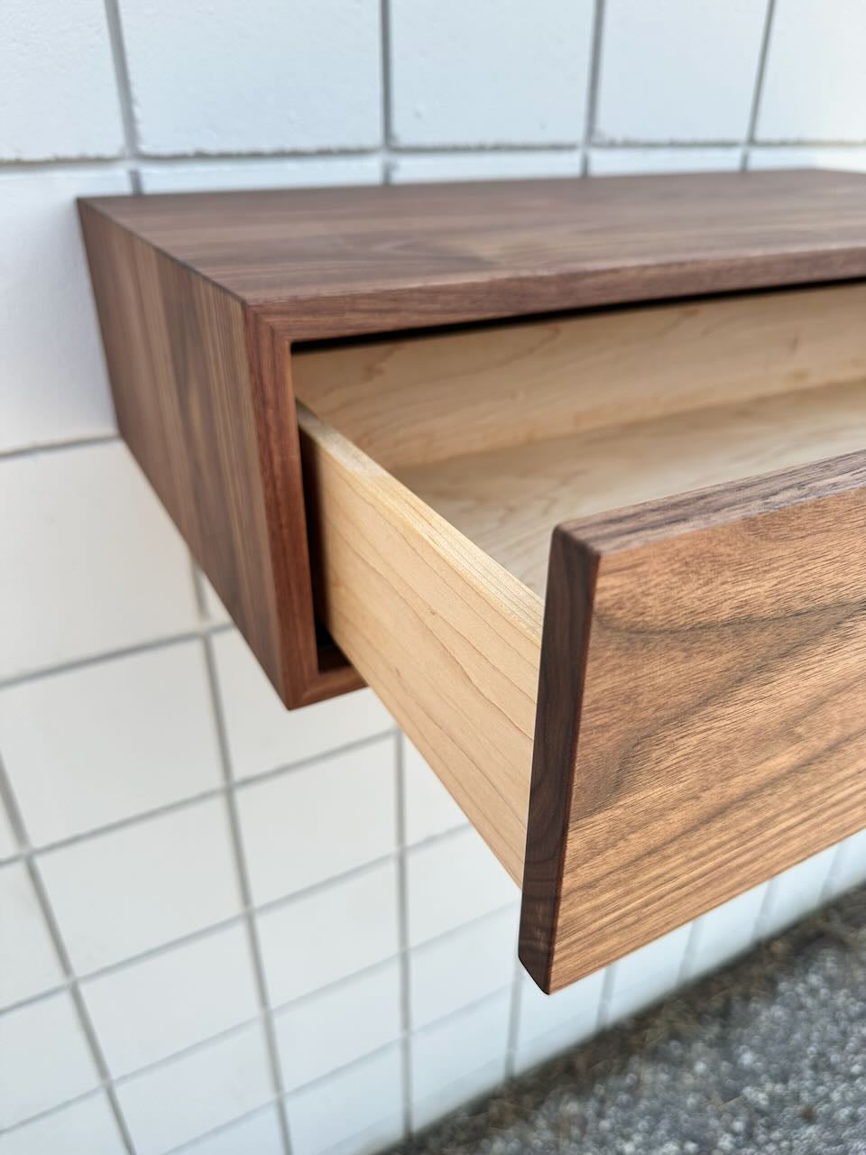 A close-up of the Krovel Furniture Co. Single Drawer Walnut Desk - SAMPLE, partially open, showcases its smooth, natural walnut finish and clean, modern lines as it is mounted against a white tiled wall.