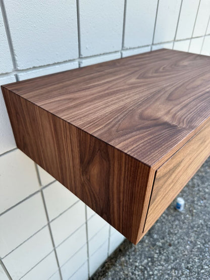 A floating wooden shelf, displaying a deep brown grain akin to the Krovel Furniture Co.’s Single Drawer Walnut Desk - SAMPLE, is mounted on a white tiled wall above a gravelly ground.