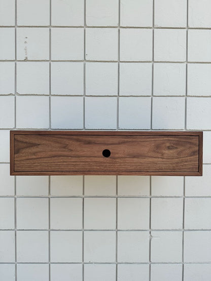 A modern and minimalistic Single Drawer Walnut Desk - SAMPLE by Krovel Furniture Co., featuring a rectangular wooden drawer with a central circular hole, is mounted on a white tiled wall with a grid pattern, evoking the look of a single-drawer desk.