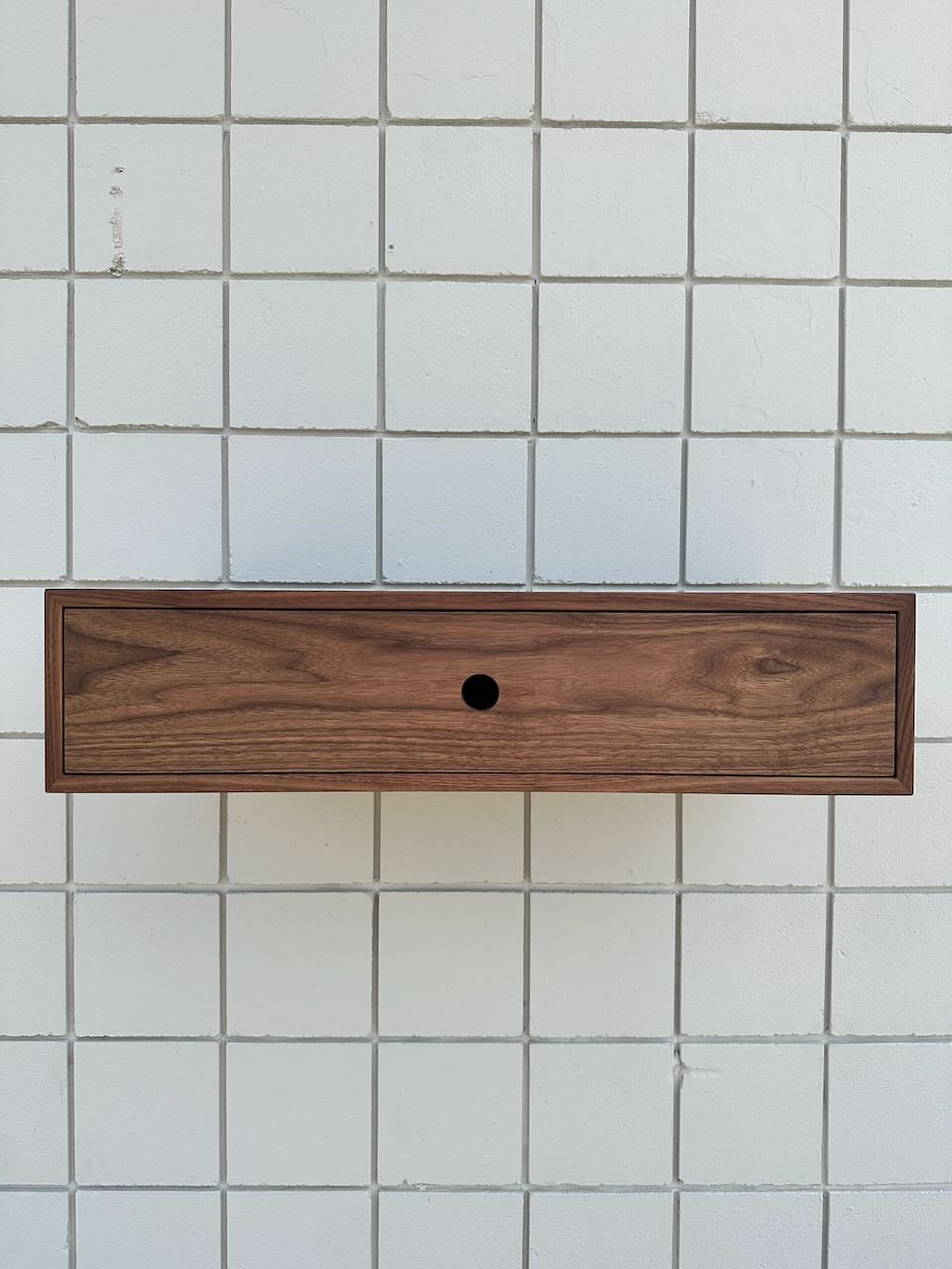 A modern and minimalistic Single Drawer Walnut Desk - SAMPLE by Krovel Furniture Co., featuring a rectangular wooden drawer with a central circular hole, is mounted on a white tiled wall with a grid pattern, evoking the look of a single-drawer desk.