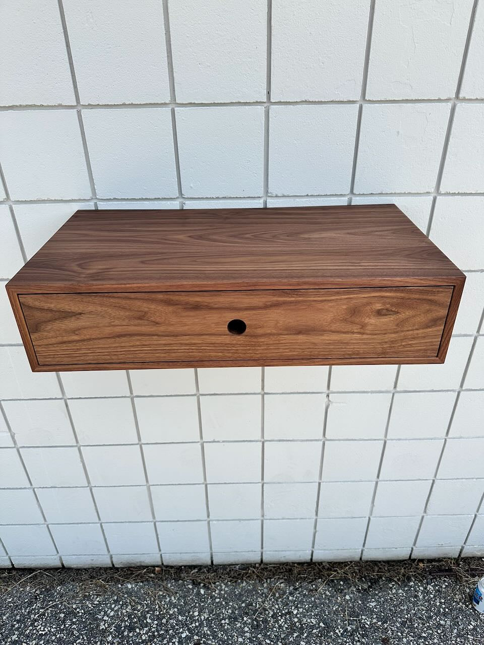 A Single Drawer Walnut Desk - SAMPLE by Krovel Furniture Co. is mounted on a white tiled wall above a gravel surface, featuring a floating wooden design with a central round pull.