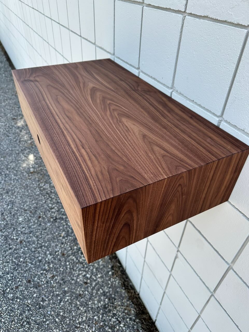 A Krovel Furniture Co. Single Drawer Walnut Desk - SAMPLE, featuring a rich wood grain reminiscent of solid hardwood, is mounted on a white tiled wall above a gravelly ground surface.