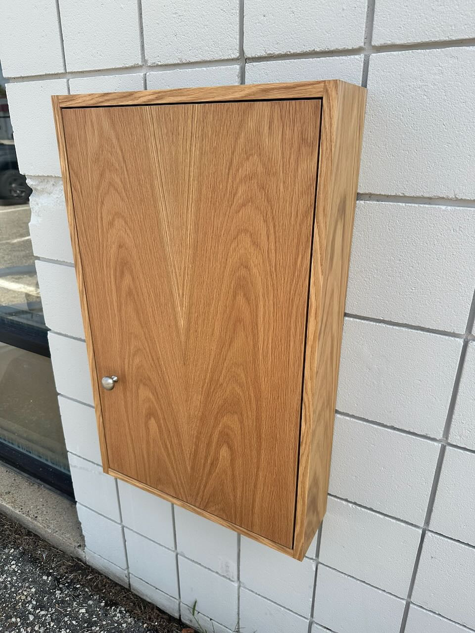 A rectangular wooden cabinet with a warm medium-brown wood grain finish and a small round silver knob, mounted against a white tiled wall.