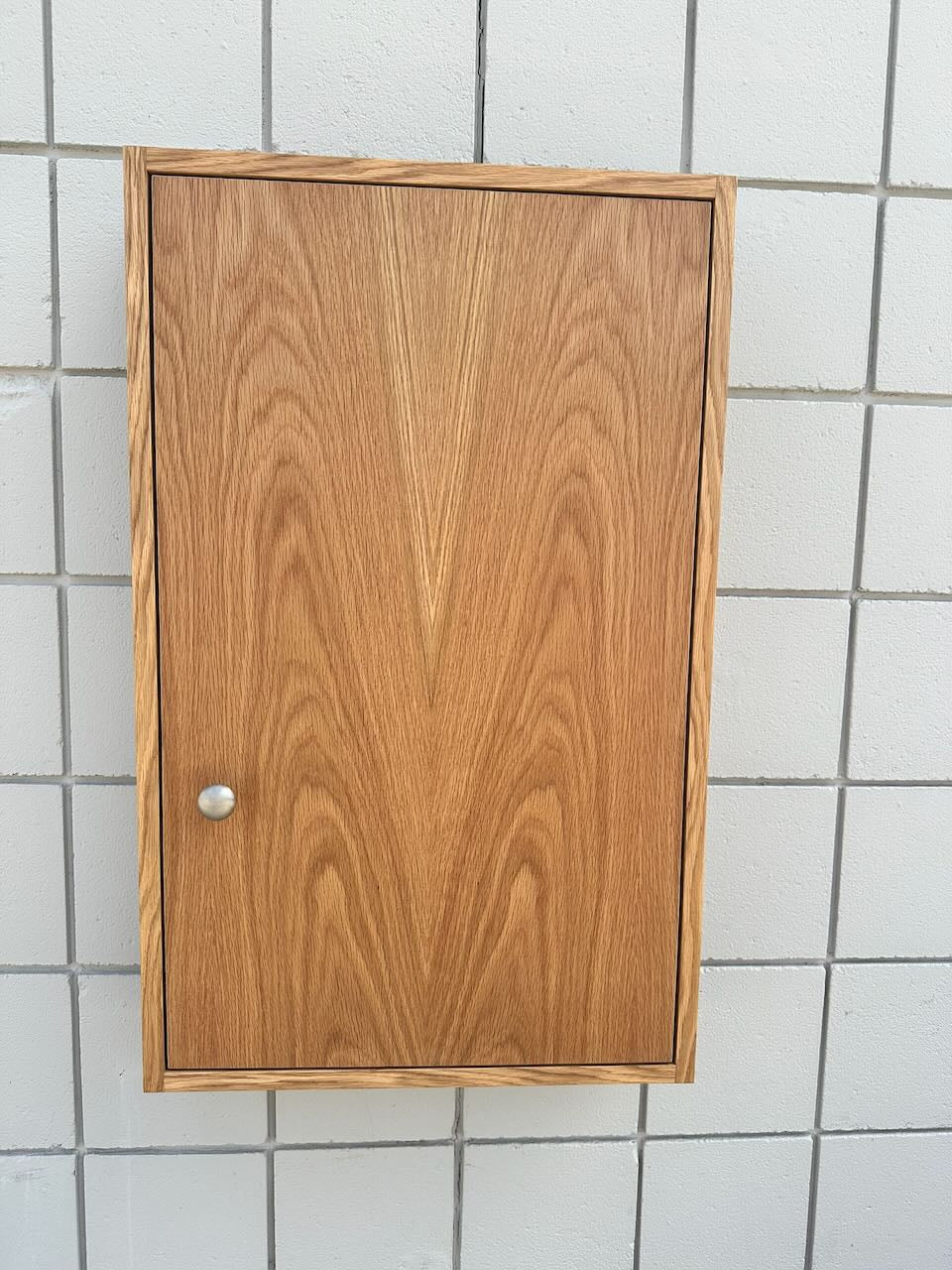 A rectangular wooden cabinet with a warm medium-brown wood grain finish and a small round silver knob, mounted against a white tiled wall.