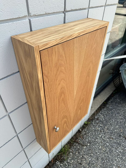 A wall-mounted wooden storage cabinet with a single door and a small round silver knob, shown attached to a white tiled exterior wall next to pavement.