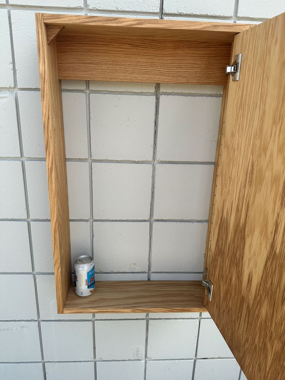 An open wooden wall-mounted cabinet with a light oak finish, shown against a white tiled wall. A small can sits on the bottom shelf inside the cabinet.