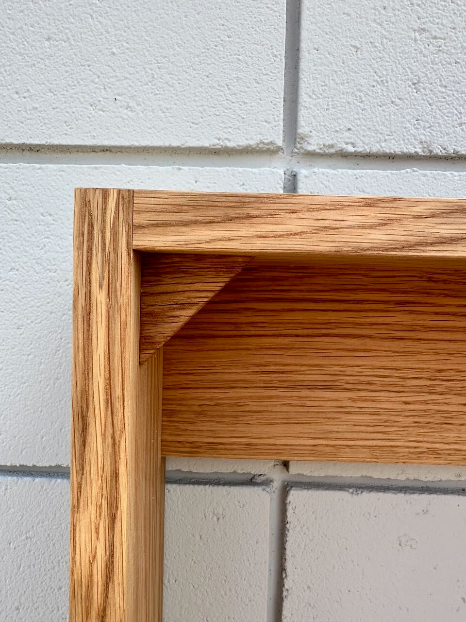 Close-up of a wooden cabinet corner with a warm medium-brown oak finish, set against a light gray painted brick wall.