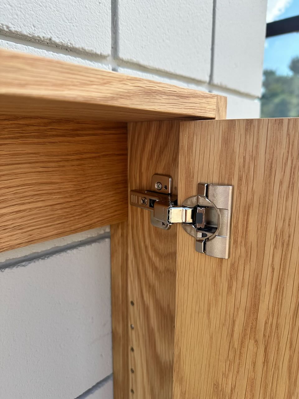 Close-up of a light oak wood cabinet door hinge attached to a matching wood cabinet, set against a white brick wall background.