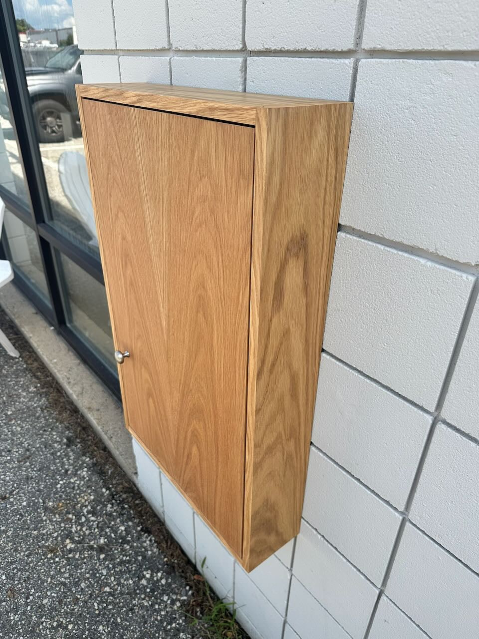 A light oak wood wall-mounted cabinet with a single door and a small silver knob, mounted on a white tiled exterior wall next to a glass window.