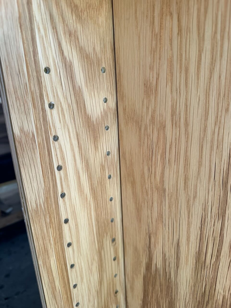 Close-up of a light oak wood cabinet door showing the wood grain texture and a row of pre-drilled screw holes along the edge.