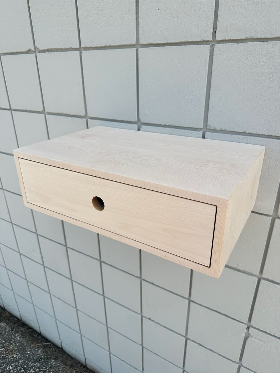 A light natural wood floating wall cabinet with a single drawer and a round cutout, mounted against a white tiled wall.