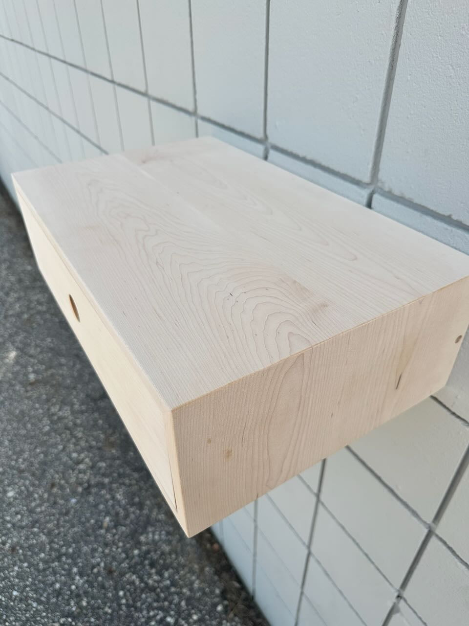 A light natural wood floating shelf mounted on a white tiled wall, shown from an angled perspective over asphalt ground.