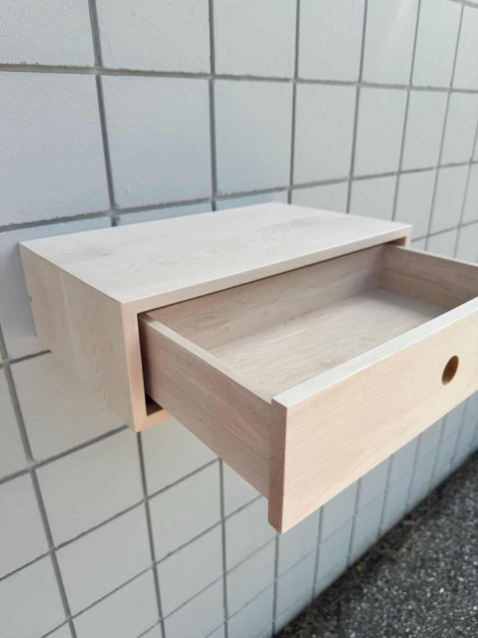 A light natural wood wall-mounted drawer with one pull-out drawer open, set against a white tiled wall.