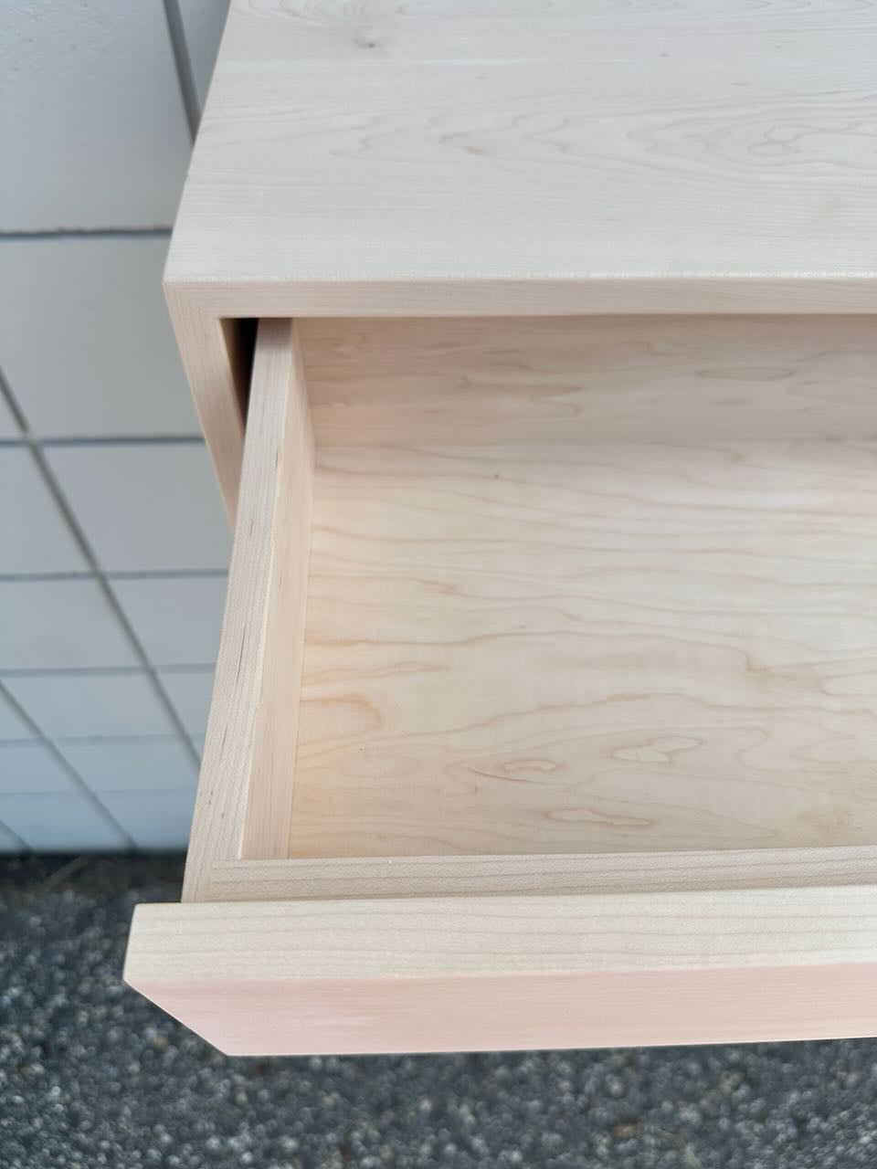 Close-up of a light natural wood drawer with one drawer pulled open, showing the wood grain and interior, set against a tiled wall and asphalt ground.