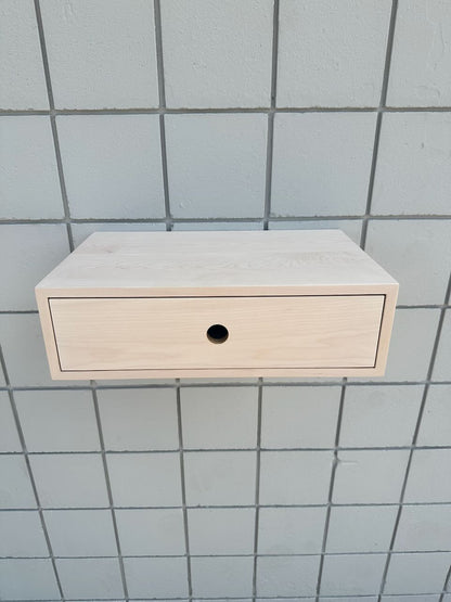 A light natural wood floating wall cabinet with a single drawer and a round black pull, mounted against a gray tiled wall.
