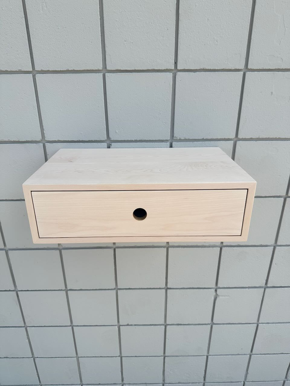 A light natural wood floating wall cabinet with a single drawer and a round black pull, mounted against a gray tiled wall.