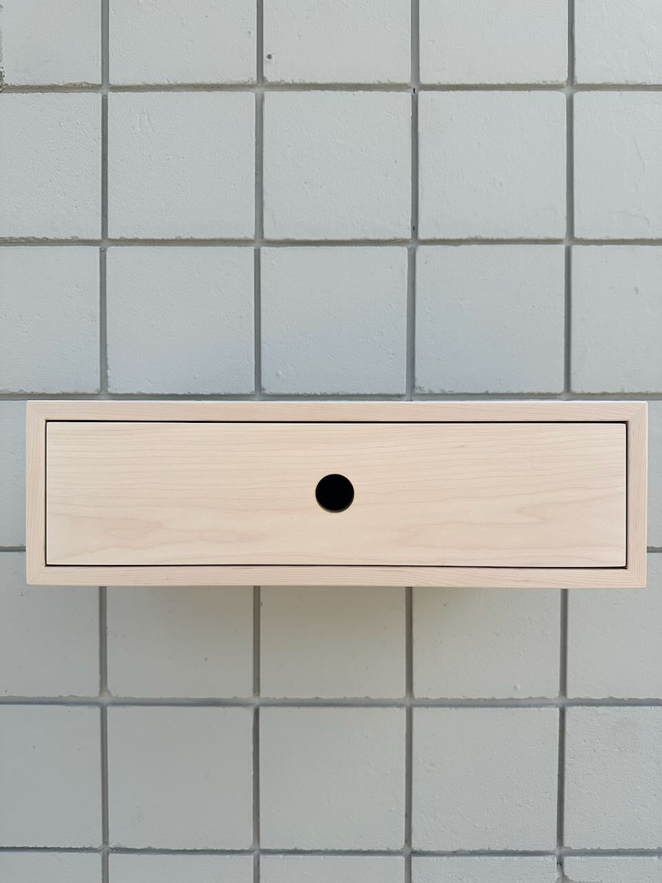 A minimalist light wood floating wall shelf with a single round black hole detail, mounted against a light gray tiled wall.
