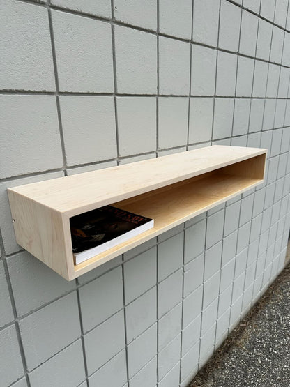 A light wooden wall shelf is mounted on a tiled wall, offering a subtle accent reminiscent of the Maple Console Table - UNFINISHED by Krovel Furniture Co. Inside the shelf are two books or magazines, with one partially visible. Below the shelf, the area remains clear, and the ground is visible at the image’s edge.