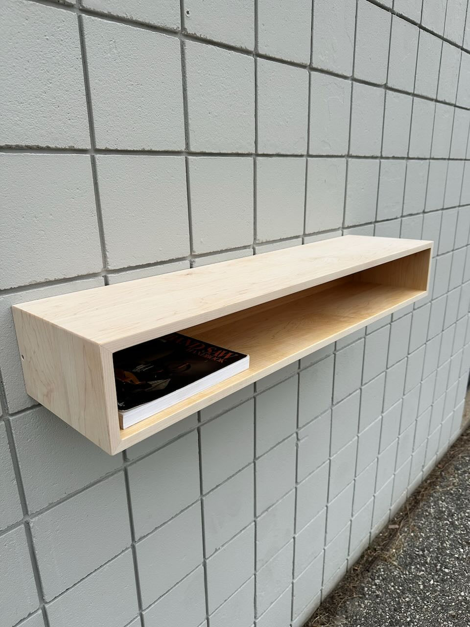 A light wooden wall shelf is mounted on a tiled wall, offering a subtle accent reminiscent of the Maple Console Table - UNFINISHED by Krovel Furniture Co. Inside the shelf are two books or magazines, with one partially visible. Below the shelf, the area remains clear, and the ground is visible at the image’s edge.