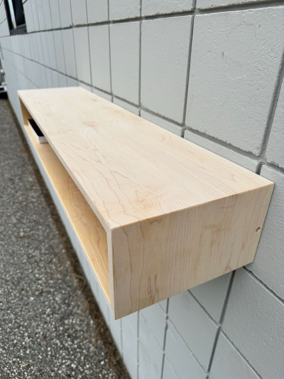 A Maple Console Table - UNFINISHED by Krovel Furniture Co. is mounted on a white tiled wall outdoors above a gravel surface, showcasing the simple elegance characteristic of discounted furniture.