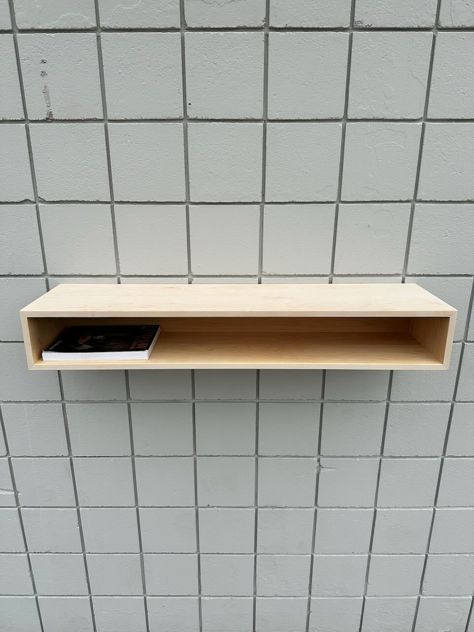 A light wooden wall shelf with an open front is mounted on a tiled wall, echoing the clean lines of Krovel Furniture Co.’s Maple Console Table – UNFINISHED. A single thin book or magazine is placed on the left side inside the shelf.