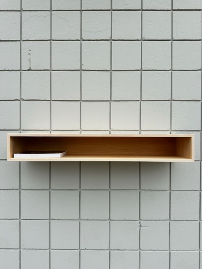 A minimalist Maple Console Table - UNFINISHED by Krovel Furniture Co. is mounted on a light gray tiled wall, holding a single closed white book placed to the left side inside the shelf.