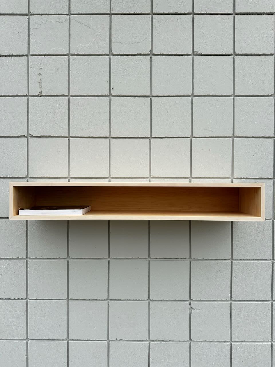 A minimalist Maple Console Table - UNFINISHED by Krovel Furniture Co. is mounted on a light gray tiled wall, holding a single closed white book placed to the left side inside the shelf.
