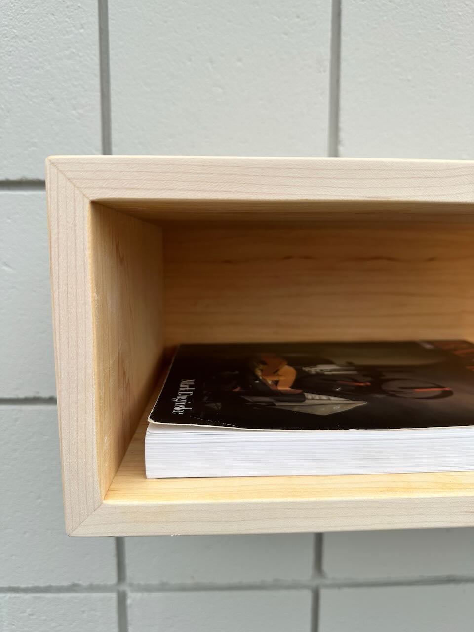 A close-up of a wooden box shelf mounted on a tiled wall. Inside the shelf sits a single paperback book with a mostly black cover depicting part of a person’s face and hands. The natural finish of the shelf echoes the clean lines of Krovel Furniture Co.’s Maple Console Table – UNFINISHED, adding warmth and character to the space.