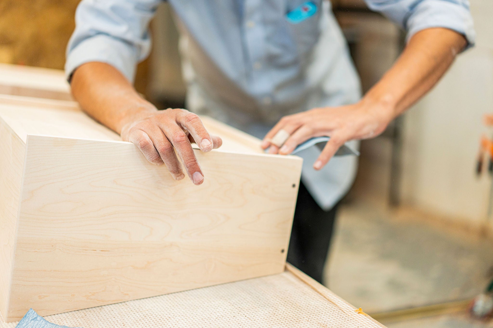 In a workshop, a person in a light blue shirt is meticulously sanding a wooden surface for Krovel Furniture Co. Their hands focus on smoothing the corner of an unfinished wood piece, emphasizing the handmade quality. Dust from this Made in USA craft is visible on their hands and the surface.