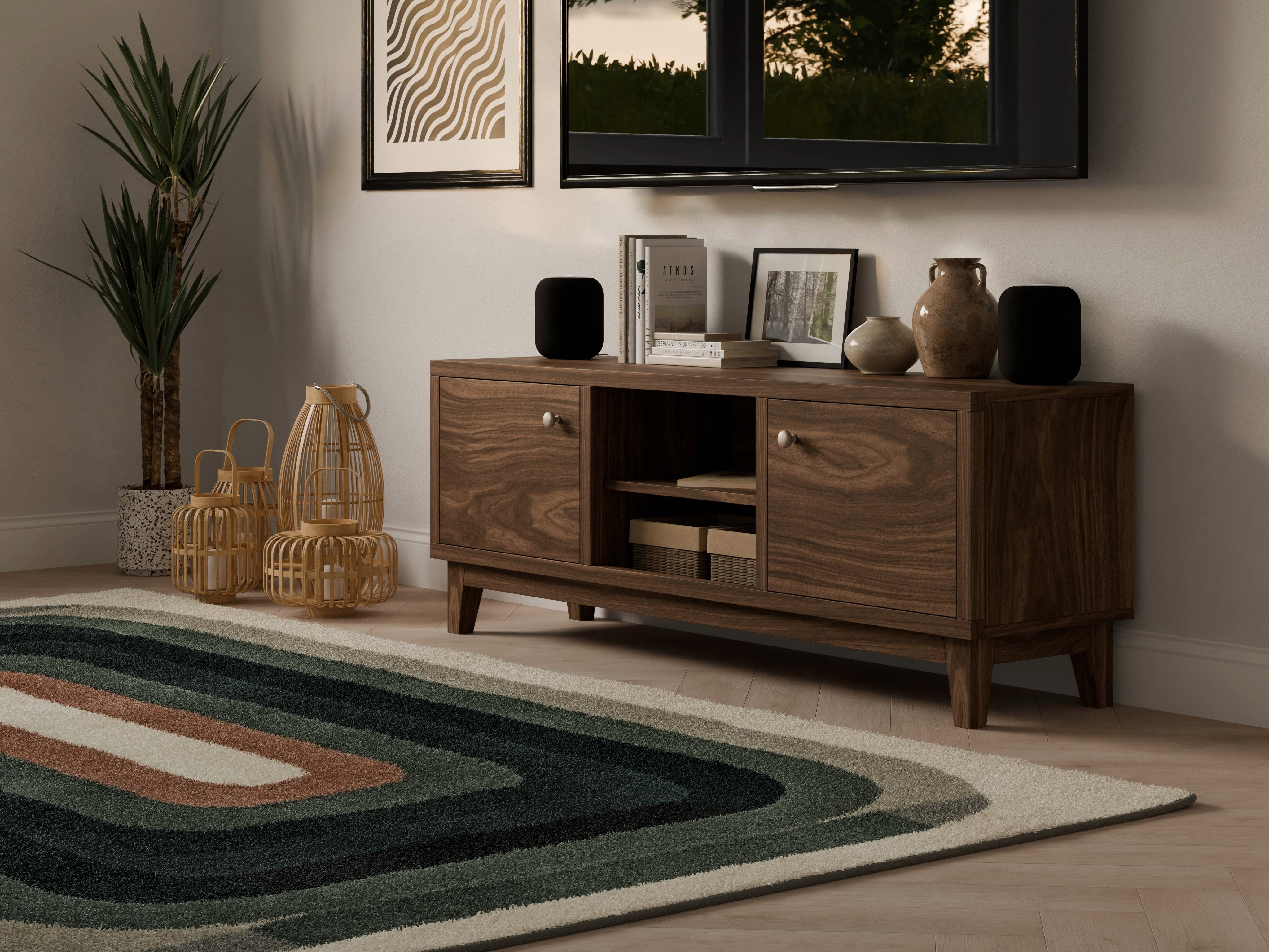 A modern living room with a wooden TV stand, books, vases, and speakers, next to woven lanterns and a potted plant. A patterned rug covers the floor, and framed art hangs on a light-colored wall.