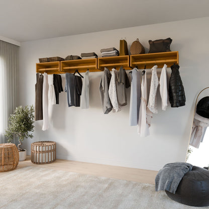 A modern, minimalistic room showcases the Closet Hanger in Cherry by Krovel Furniture Co., wall-mounted with assorted neutral garments. A soft beige rug covers the floor beneath. A round mirror rests against the wall next to a small plant and woven baskets.