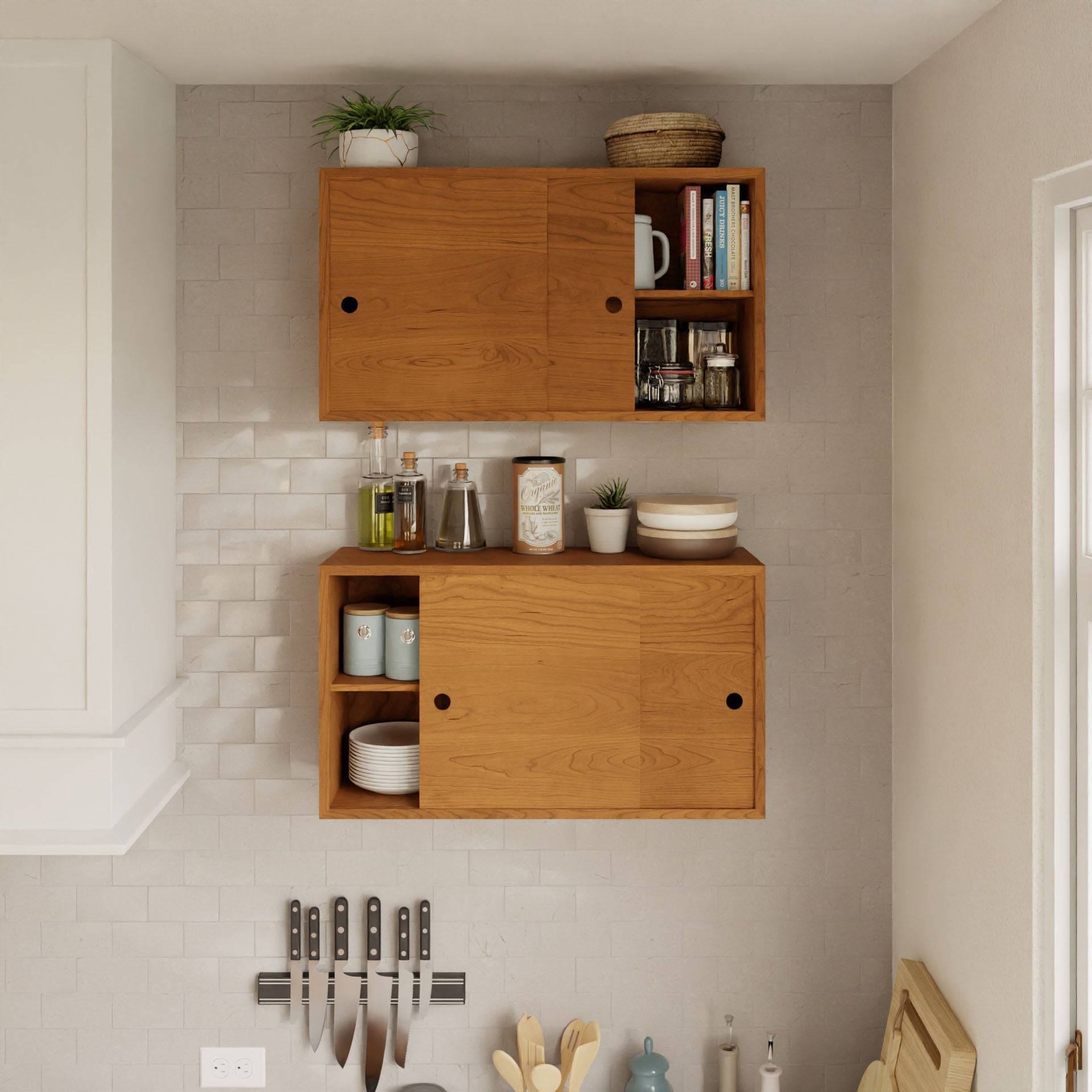 Krovel's Cupboard with Shelf in Cherry is mounted on a textured white wall, featuring sliding doors. The top shelf holds a plant, a mug, and books, while the lower one stores cups and condiments. A magnetic strip with knives underneath completes this elegant setup.