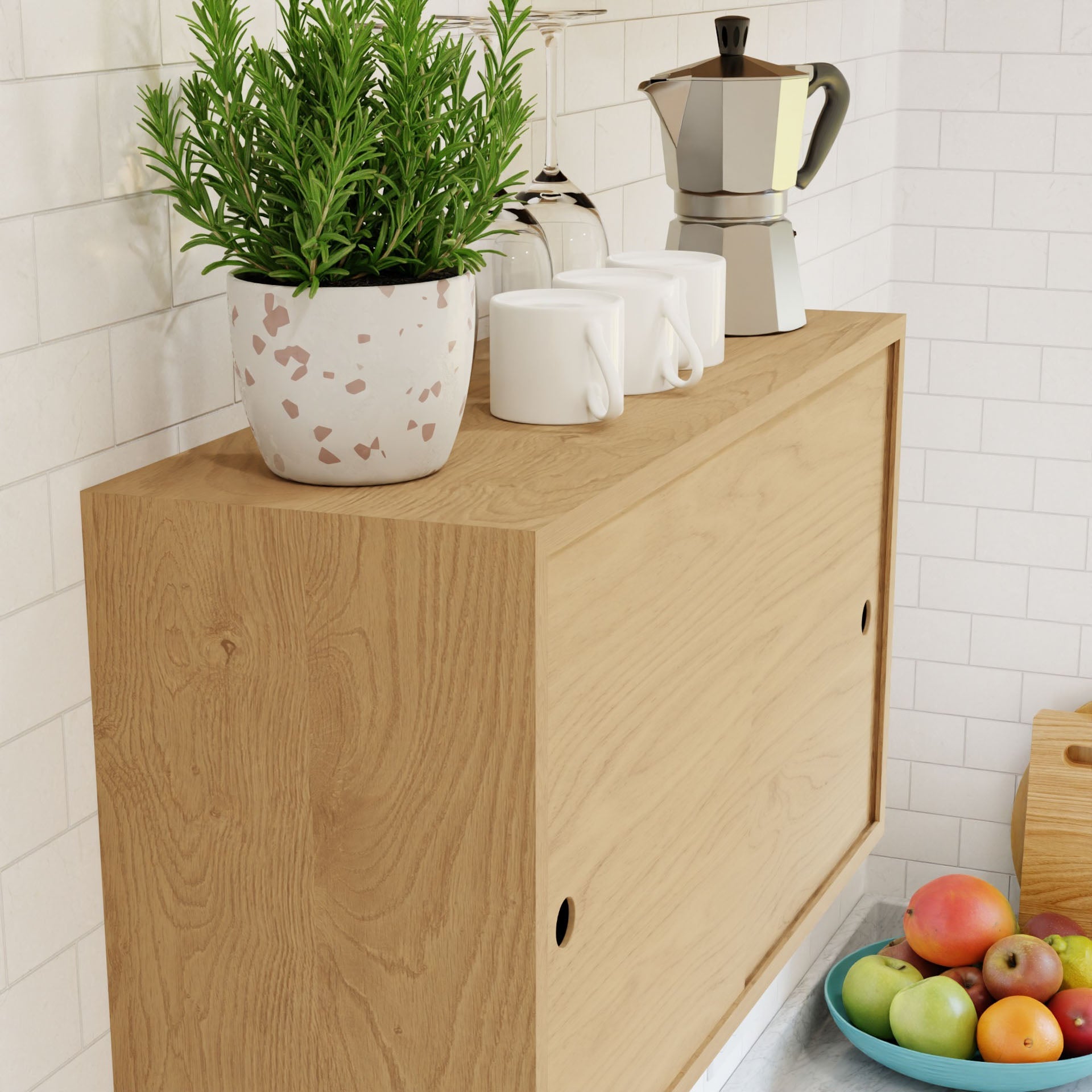 A Krovel Cupboard with Shelf in Maple holds a potted plant, three white mugs, and a silver moka pot against white tiled walls. A basket of colorful fruits, including apples and oranges, is partially visible beneath the sleek walnut finish.